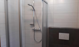 A modern shower with a glass partition and a handheld showerhead. The walls are light-tiled and the floor is dark.