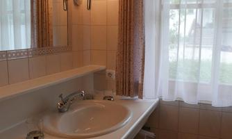 A bright bathroom with a sink and a large mirror. The windows are decorated with white curtains, and the room is designed with beige tiles.