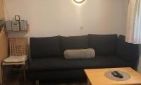 A cozy living room with a dark gray sofa and a wooden table. There is a lamp on the wall and a chair stands opposite.