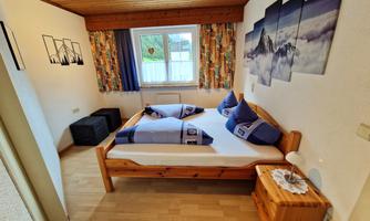A cozy bedroom with a wooden bed and blue cushions. The walls are decorated with pictures and colorful curtains.