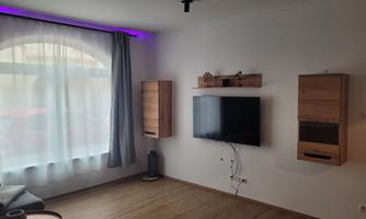 A modern living room with a large window that lets in plenty of natural light. Two wooden shelves and a television hang on the wall.