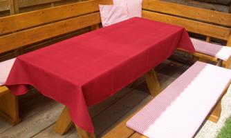 A wooden banquet table with a red tablecloth is set up in an outdoor area. The benches are covered with cushions in a checkered pattern.