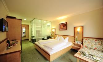 A modern hotel room with a double bed and a cozy seating area. The bathroom is separated from the room by glass walls.