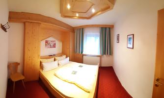 A cozy bedroom with a double bed and wooden furniture. There is carpet flooring and windows with curtains.