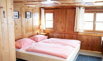 A cozy bedroom with wooden-paneled walls and a double bed decorated with red pillows. There are windows with curtains and a small lamp on a bedside table.