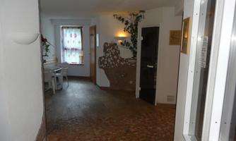 A bright hallway with a patterned floor and a small seating area. Decorative elements and plants can be seen on the walls.