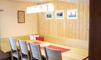 A bright dining area with a large wooden table and six chairs. Pictures hang on the wall and a modern lamp illuminates the space.