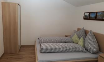 A cozy bedroom with a large bed and checkered pillows. The room has bright walls and a wooden floor.