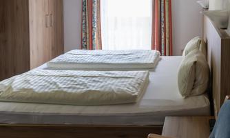 A simple bedroom with a double bed, two mattresses, and a comfortable chair. The walls are light-colored, and there are windows with curtains.