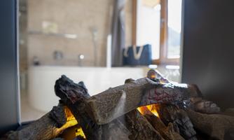 A fireplace with burning logs in the foreground. In the background, a bright bathroom with a bathtub and window can be seen.