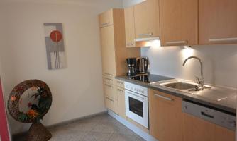 A modern kitchen with light wood cabinets and a stainless steel sink. Next to the kitchen is a decorative mirror with a plant arrangement.