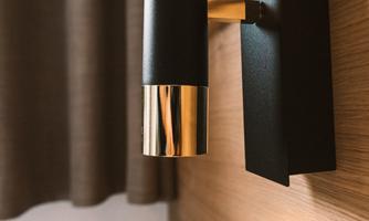 A modern wall lamp with a black and gold design. In the background, a light curtain and a wooden surface are visible.