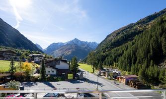 A picturesque alpine landscape with snow-covered mountains and green forests. In the foreground, some buildings and a quiet road can be seen.