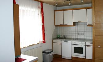 A modern kitchen with white cabinets and a bright countertop. The room has large windows with red curtains and a wooden floor.