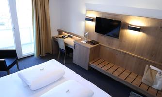 A modern hotel room with a double bed, a desk, and a television. Large windows provide plenty of daylight.