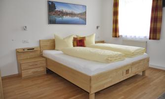A bright bedroom with a large double bed and wooden shelves. On the wall hangs a picture of a landscape with water and trees.