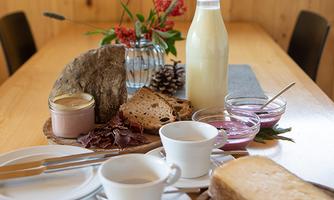 A rustic breakfast table with fresh bread, spreads, and a bottle of milk. Cups and a decorative plant are on a wooden table.