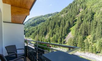A balcony with two chairs that offers a view of green mountains and dense forests. The landscape is sunny and inviting.