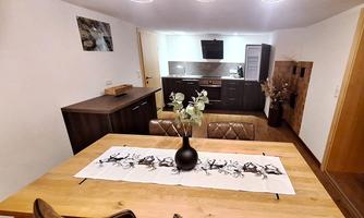 A modern dining room with a large wooden table and brown chairs. The walls are light, and the kitchen in the background is elegantly and functionally designed.