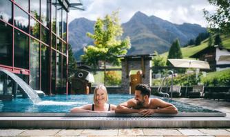 A couple relaxes in the outdoor hot tub with a view of the mountains. The surroundings are green and inviting, in nice weather.