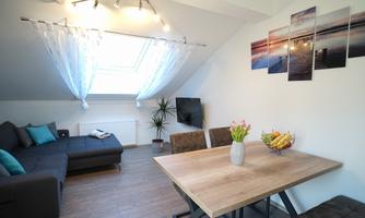 A modern living room with a comfortable couch and a dining table. Pictures of a landscape hang on the wall, and there is a small plant.