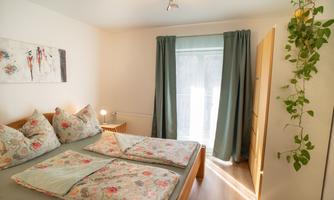 A cozy bedroom with a large bed and colorful pillows. On the table, there is a lamp and the room has large windows with curtains.