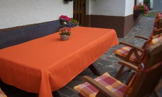 A table with an orange tablecloth stands on a stone floor. Around the table, there are wooden chairs with checkered cushions and colorful flowers are decoratively arranged.