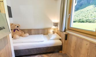 A cozy bedroom with a single bed and a large plush bear. The windows offer a beautiful view of the green landscape.