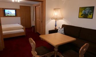 A cozy room with a wooden table and two chairs. In the background, there is a bed and a TV.