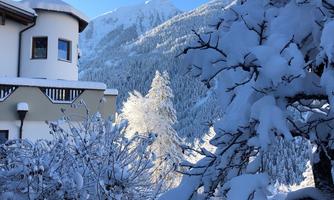 A snow-covered landscape with a cozy house and snow-covered trees. In the background, majestic mountains and a blue sky can be seen.