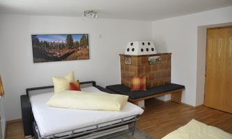 A cozy bedroom with a sofa bed and decorative pillows. The wall has a picture of a mountain landscape and there is a tiled stove.