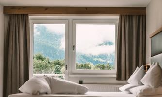 A modern bedroom with a large window and a view of the mountains. The room is bright and inviting, with cozy cushions on the bed.