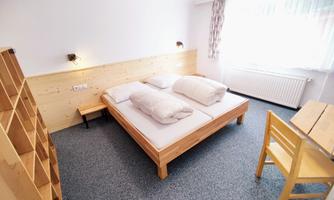 A cozy bedroom with a large double bed and two pillows. The room is bright and simply furnished, with a wooden table and chair.