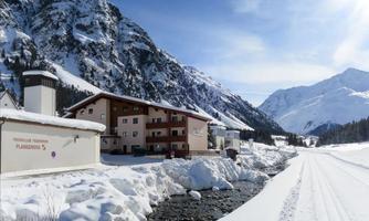 A quiet winter landscape with snow-covered mountains. Nearby, a clear stream flows beside a cozy building.