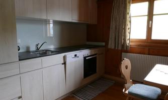 A modern kitchen with wooden cabinets, a sink, and an oven. A table with a chair is next to a window that lets in daylight.