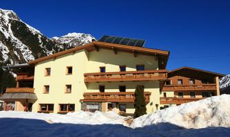 A sunny building in the mountains with several balconies. Snow lies in front of the house, and the sky is clear and blue.