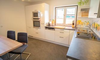 A modern kitchen with white cabinets and high-quality appliances. Four chairs are at the table, and a large window provides plenty of light.