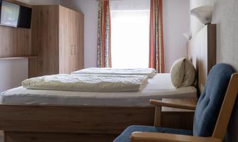 A simple room with two single beds, a chair, and a television. Bright curtains let in a lot of daylight.