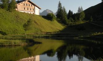 A picturesque house sits by a tranquil lake, surrounded by gentle meadows and trees. In the background, a majestic mountain rises.