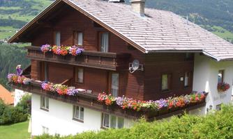 A picturesque house with a wooden facade and colorful flowerboxes on the balcony. The surroundings are surrounded by green meadows and mountains.