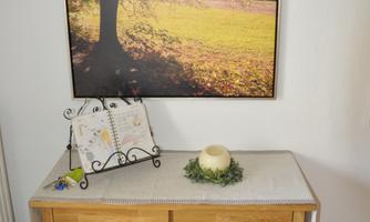 A wooden cabinet with a tabletop and a decorative candle stands in front of a wall with a large picture of a tree in sunlight. Next to it, a calendar lies on a small stand.