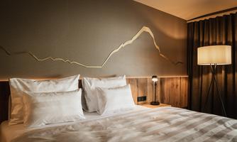 A modern bedroom with a padded bed and elegant lighting. The wall features a stylized mountain motif and is framed with cozy curtains.