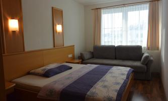 A simple room with a double bed and a gray sofa. Large windows provide plenty of daylight.