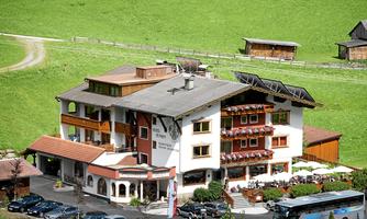 A charming hotel in a green landscape, surrounded by meadows and mountains. The buildings have balconies with blooming flowers and are surrounded by cars and a bus.