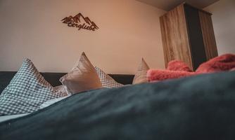 A cozy bedroom with decorative cushions and a wooden piece of furniture. A wooden sign hangs on the wall with the word "Mountain Happiness."