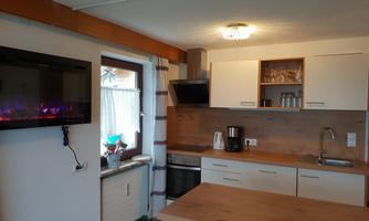 A modern kitchen with a wooden workspace and white cabinets. The room contains a television and a coffee machine.