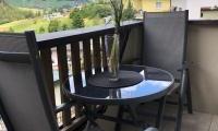 A balcony with a round table and two chairs. A vase with a flower is on the table, while a green landscape can be seen in the background.