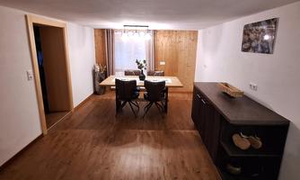 A modern dining room with a large table and four chairs. The room features warm wooden design and pleasant lighting.