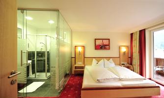 A modern hotel room with a double bed and an adjoining glass bathroom. The decor is bright and inviting with carpeted floors and warm colors.