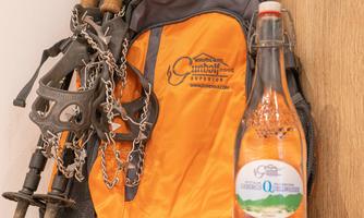 An orange backpack with hiking poles and a bottle of water is standing next to two apples. The background is made of wood and conveys a natural atmosphere.